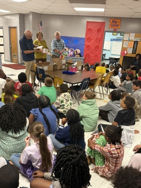 Two men standing in front of a classroom with many seated children. One man holds a book.
