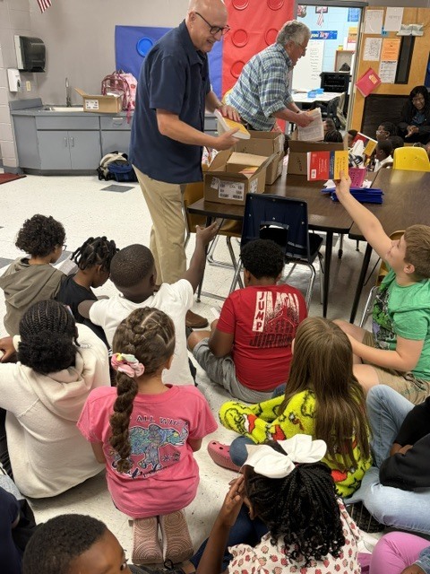 Classroom with kids sitting in a circle. A man stands behind a table with boxes on it.