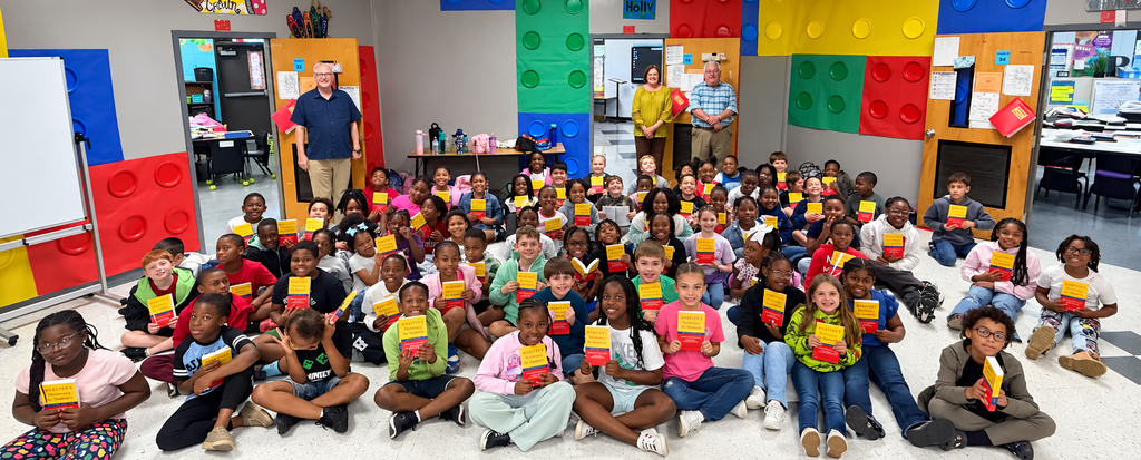 A group of children sitting in rows holding yellow blocks, with adults standing in the background.