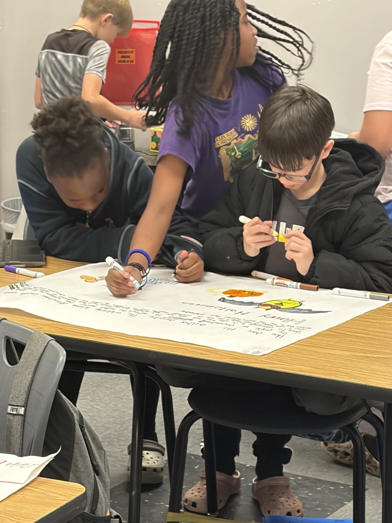 Four children at a table, three of them are looking at a large piece of paper on the table.