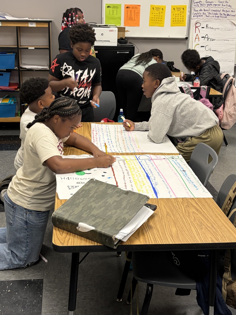 Four students work at a table, with papers and pens. One uses a pencil. There are backpacks on a chair.