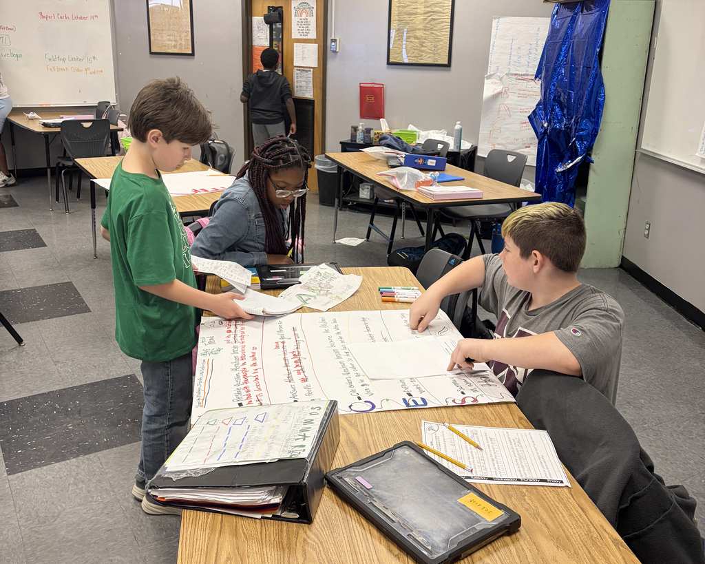 Two students in a classroom reviewing papers at a table while a teacher stands behind them.