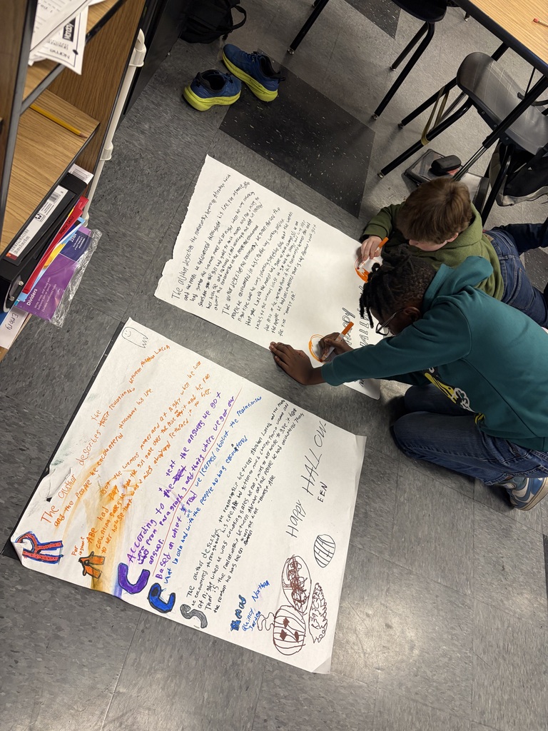 Two students are on the floor, one writing on a large white sheet of paper. The paper has various words and images in multiple colors. A wooden shelf is in the background.