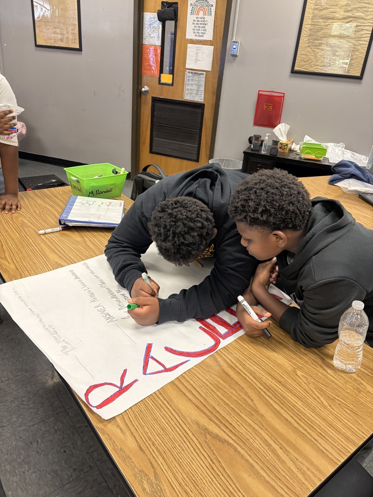 Two students collaborate on a large poster at a table, using markers. A door and bulletin board are in the background.