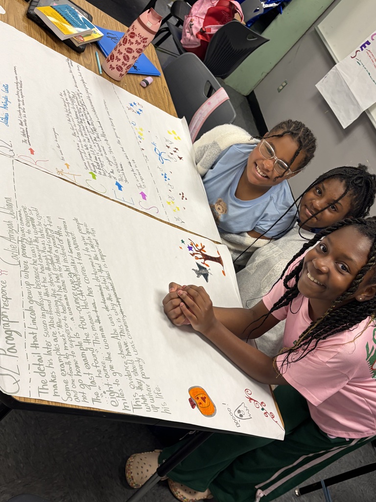 Three students sit at a table with a large sheet of paper displaying colorful illustrations and text.