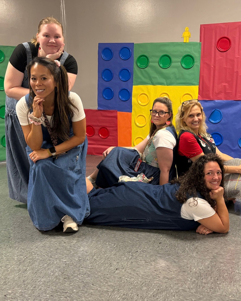 Five women in denim overalls pose for a picture in front of a backdrop of colorful Lego blocks.