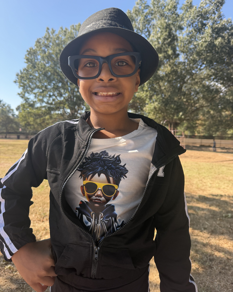 A child smiles, wearing a hat, glasses, and a jacket. The jacket has a graphic of a person with sunglasses. Behind the child, grass and trees are visible.
