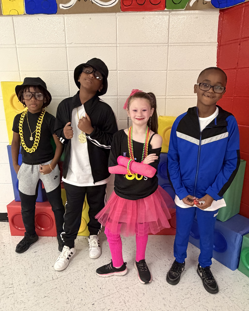 Four children pose in front of multicolored building blocks; three boys and a girl. One wears a hat and necklace.