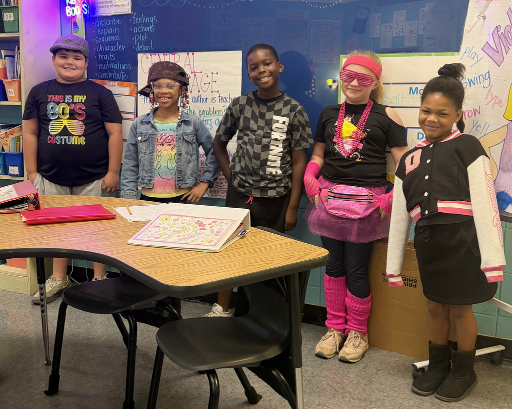 Five young students, with hats and glasses, stand in a classroom. They are near a table with papers and books.