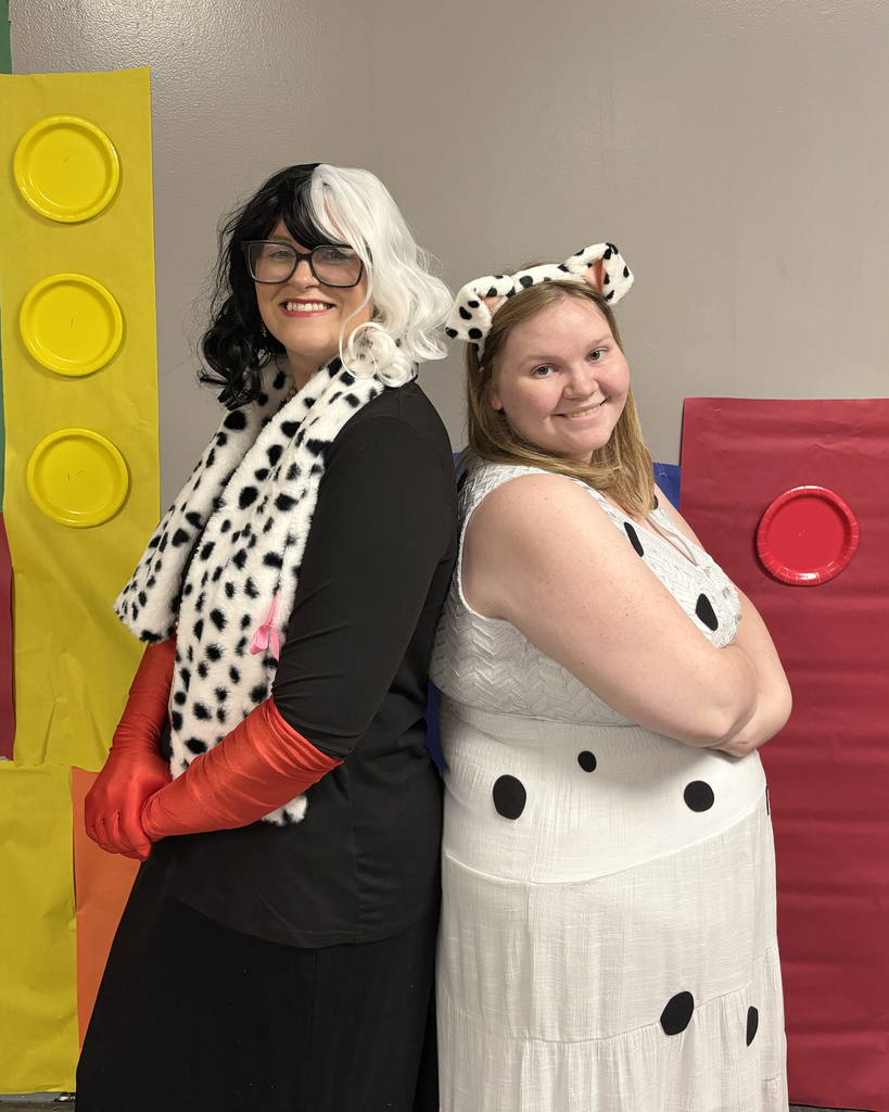 Two women in costume stand in front of a backdrop with yellow, red, and blue Lego blocks.
