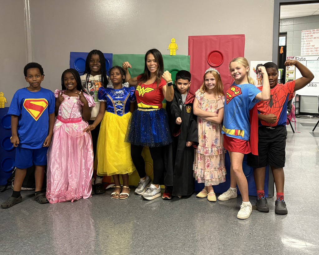 Kids in costumes, one with a Superman cape, pose for a group photo in a room with a colorful backdrop.