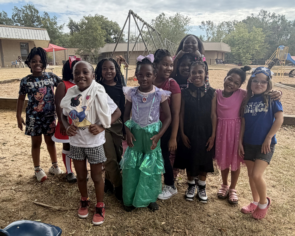 Several young girls in various outfits pose in a dirt field in a park with trees and playground equipment.