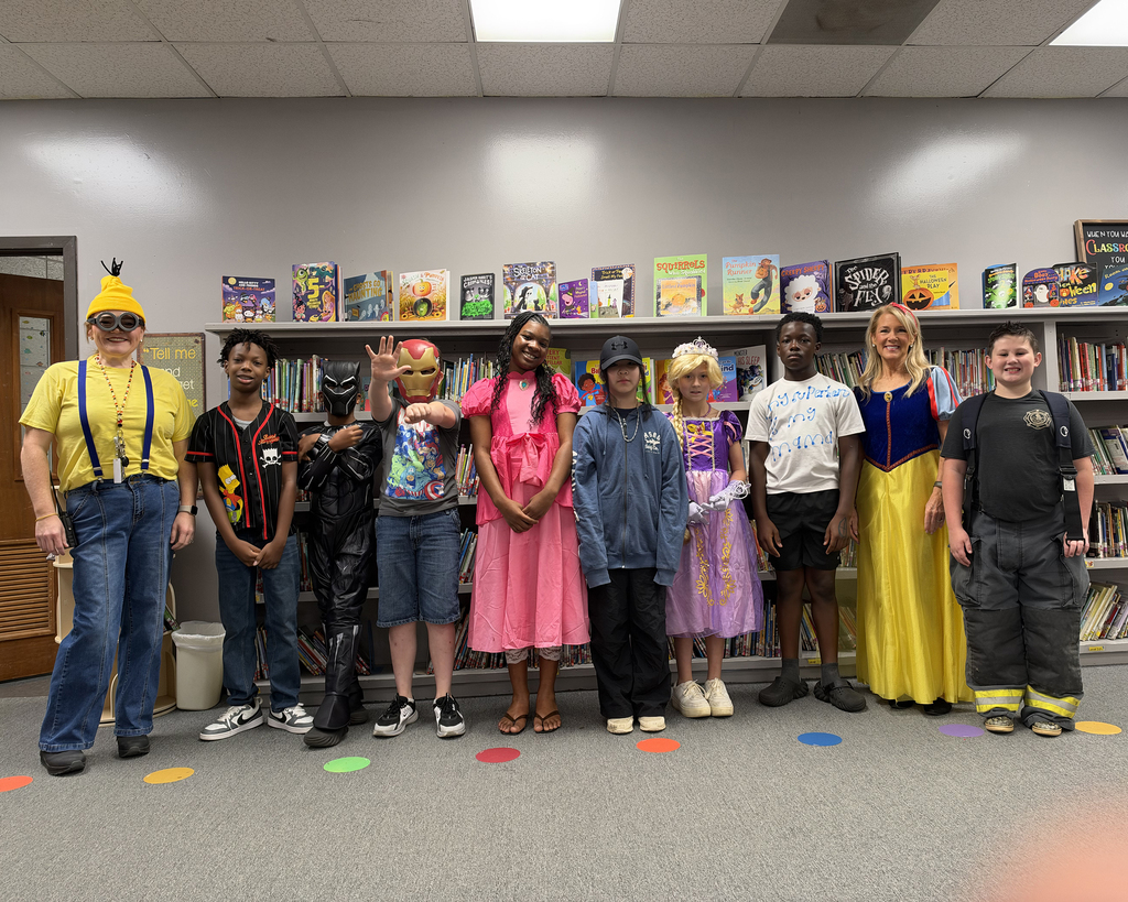 Group of students in costumes standing in a library, some with face masks, colorful circles on floor.