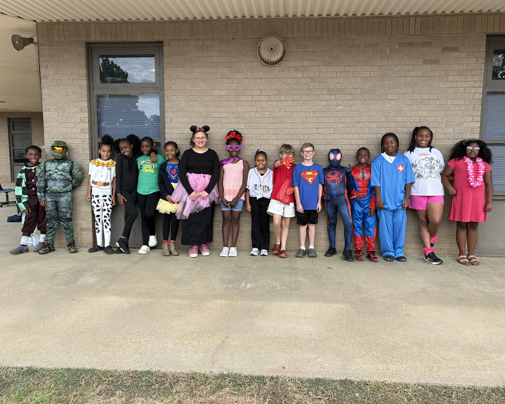 Kids in costumes stand in line outside a brick building with windows and a clock.