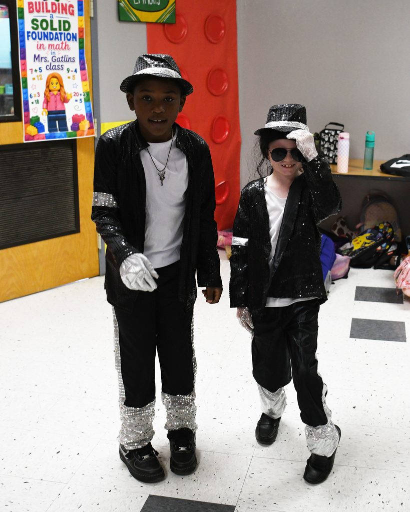Two children in matching black costumes with hats and gloves, standing in a room with a sign reading "BUILDING SOLID FOUNDATION".