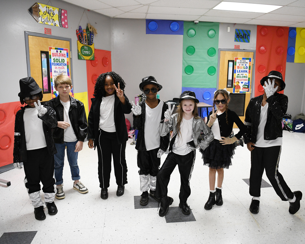 Group of children dressed in black and white, with hats, posing for a photo in a colorful classroom.