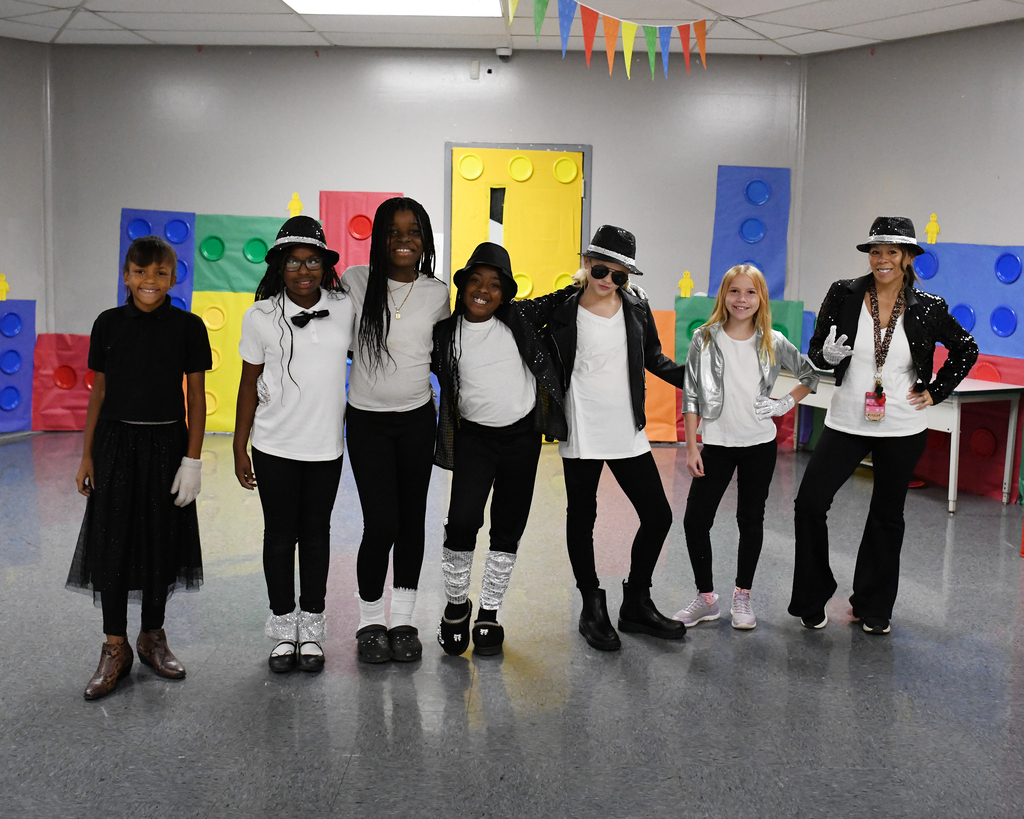 Seven individuals dressed in black and white outfits, hats, and sunglasses pose in a room with colorful Lego walls.