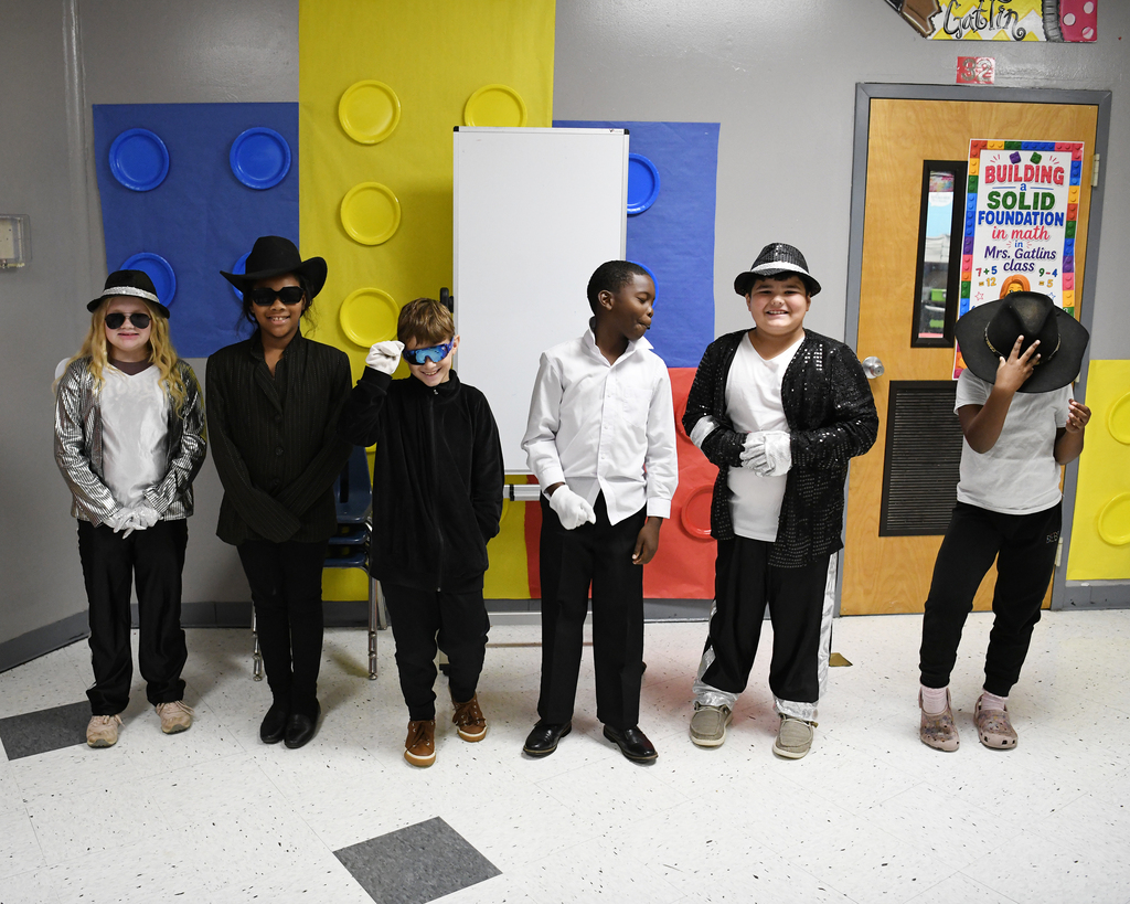 A group of children in matching outfits, hats, and sunglasses standing in a room with colorful blocks and a door.