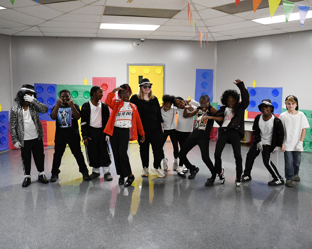 A group of young people and a woman are dancing in a room with colorful blocks and ceiling lights.