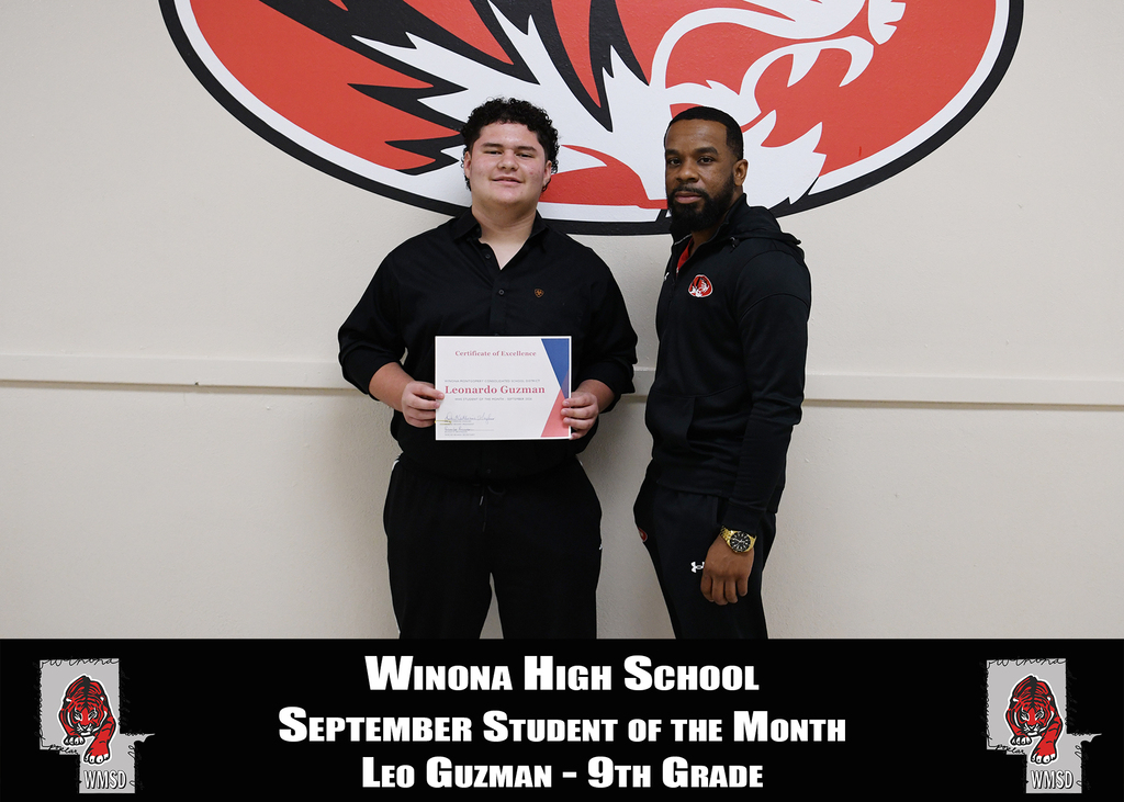 Two men stand in front of a white wall with a red school logo. They hold a certificate; one is a student, and the other is a teacher.