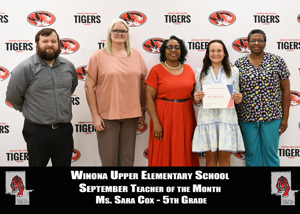 A group of five adults stands in front of a wall, with a "Tigers" logo. A woman in red holds a certificate.