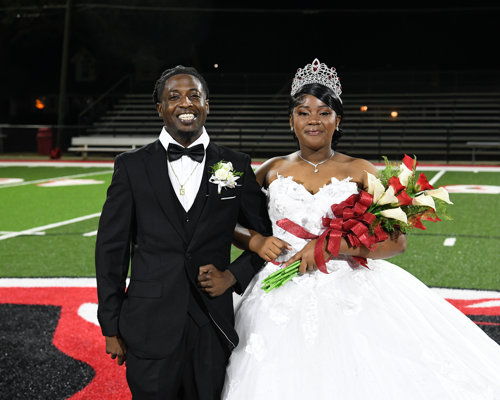 Couple in formal wear, man in tuxedo and woman in ball gown, holding flowers, on a football field.