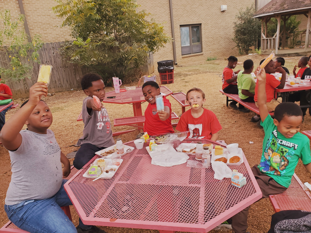 Children at outdoor tables hold up popsicles. A table has plates of food, condiments, and paper.