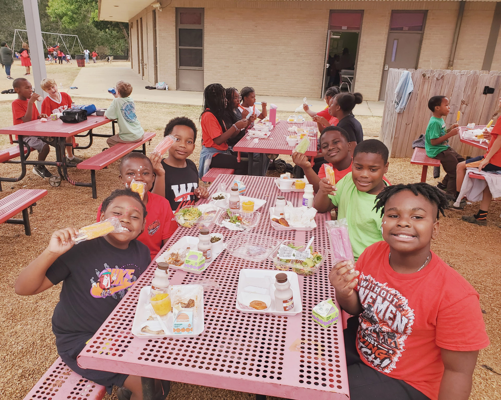 Students seated at tables outside a school, eating food. Some hold ice pops. Background shows a building, other tables, and people.
