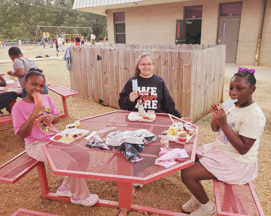 Three children eating at a table in a school courtyard. One has a popsicle, and two have food on plates.