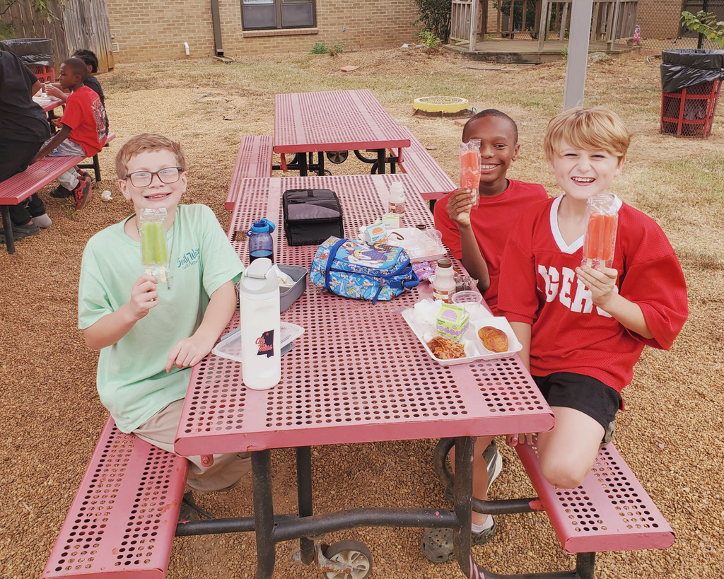 Three children sitting at a table with a red surface holding a popsicle. One child holds a blue backpack.