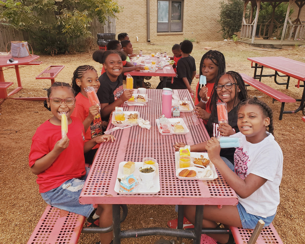 Several children eating outdoors at a picnic table, some holding ice pops. A building and trees in the background.