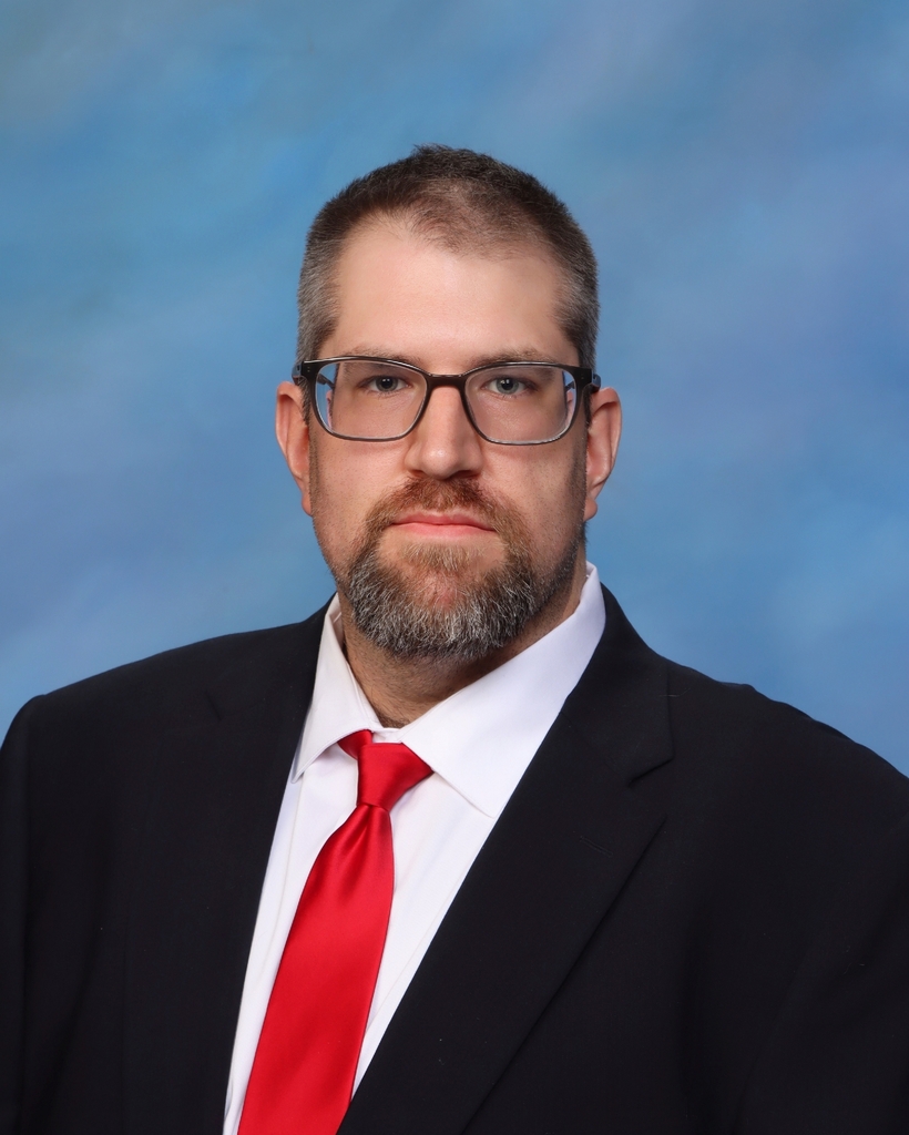 A man with a beard and mustache wearing a black suit and red tie looks at the camera.