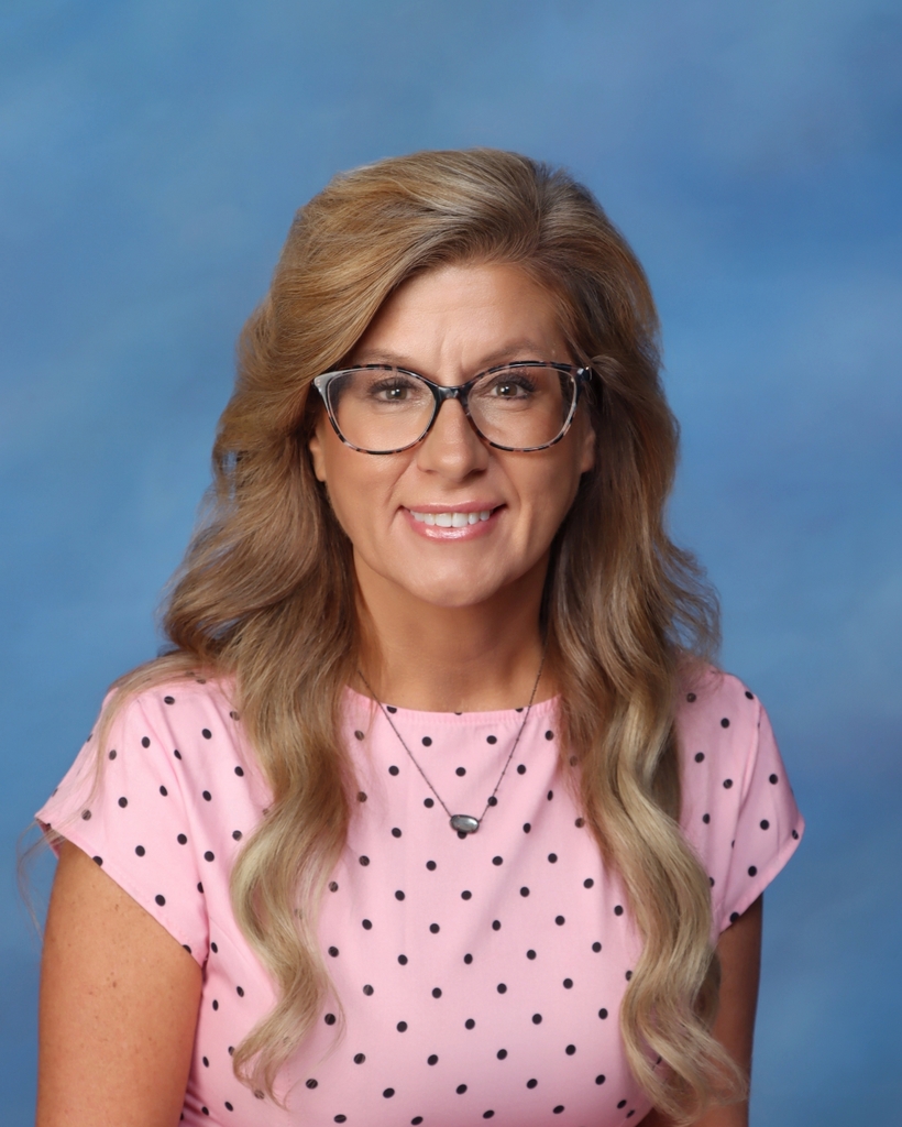 Woman with blonde hair, wearing glasses and a pink shirt with black dots, smiling at the camera.