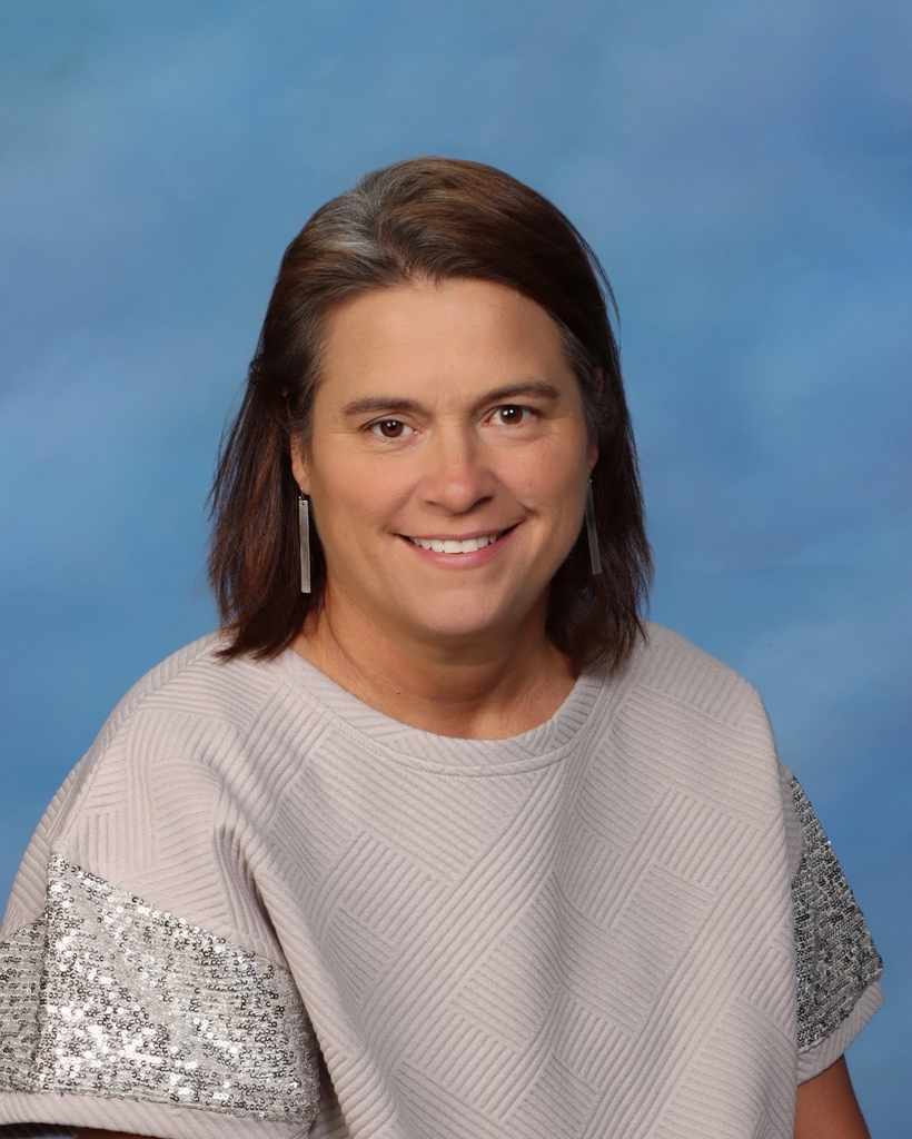 Woman with short, brown hair smiles at camera. She wears a light-colored blouse with silver, sparkly shoulders.