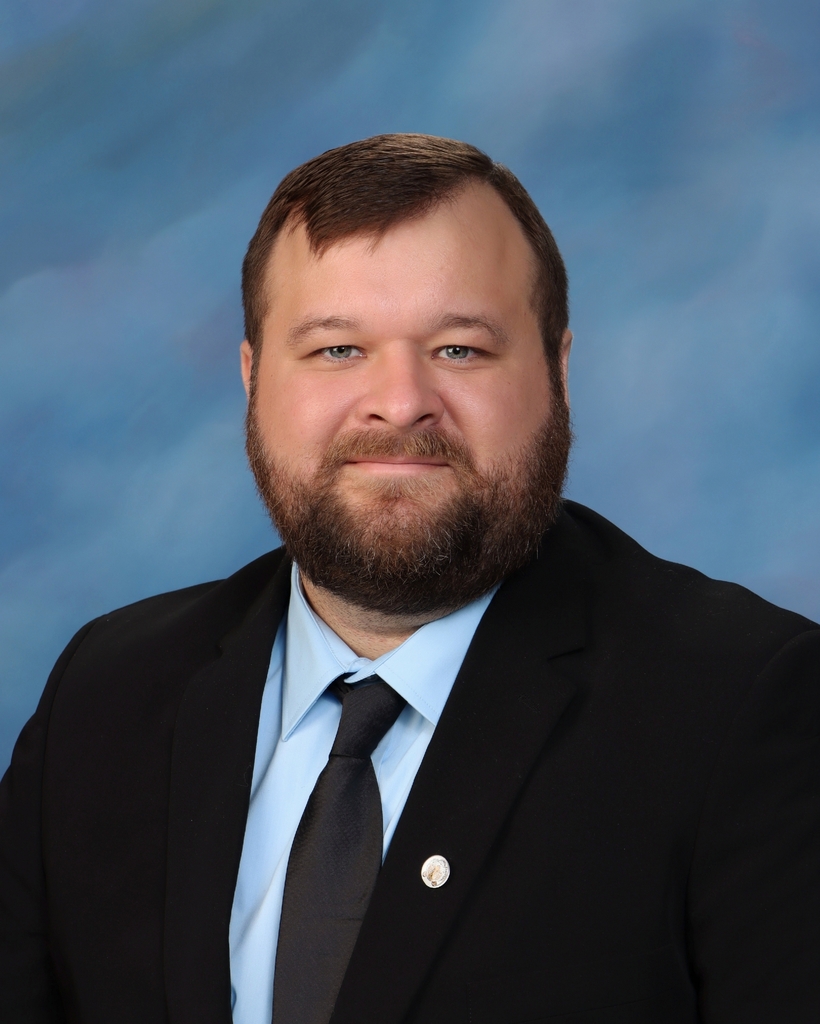 Man wearing black suit jacket, dark blue tie, and light blue shirt smiles against blue background.