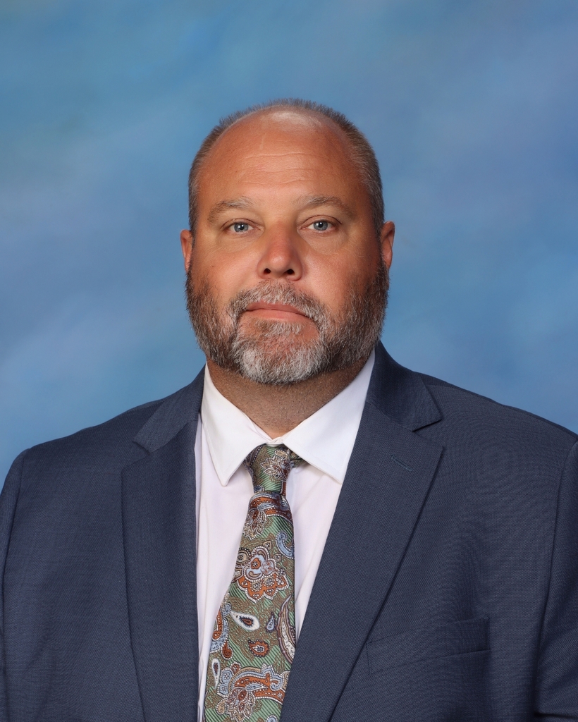 A man with a beard in a suit and tie looks at the camera, with blue background.