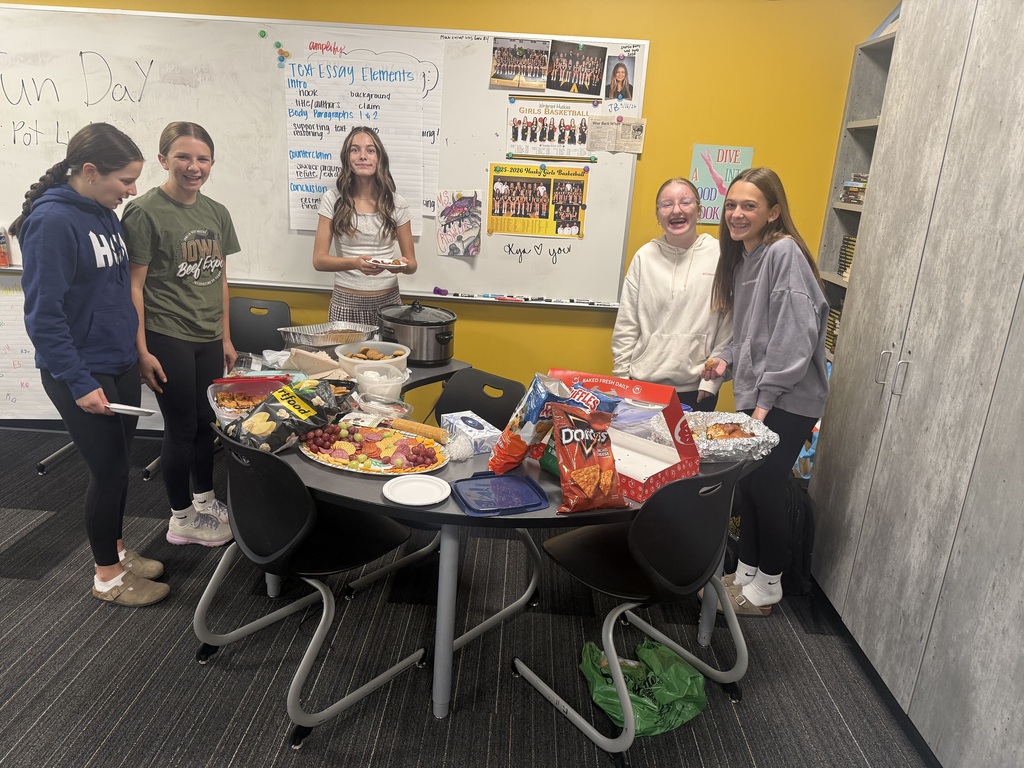 Five individuals surround a table with various foods. They stand near a whiteboard. The room has a yellow wall.