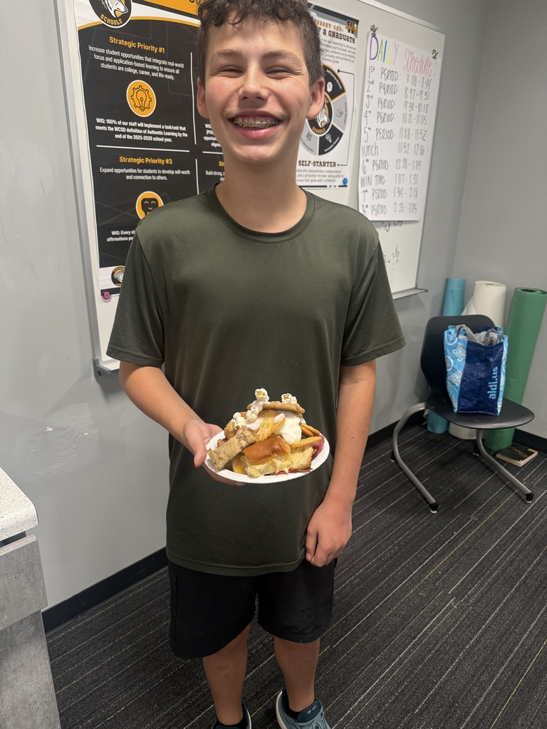 A person wearing a green shirt and black shorts holds a plate of food in a room with a white wall and a poster.