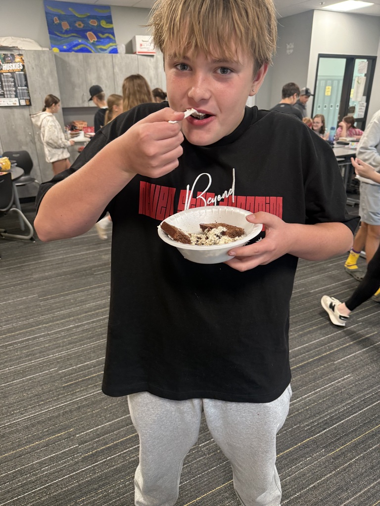 A boy eats dessert from a white bowl, wearing a black t-shirt. Several people are in the background.
