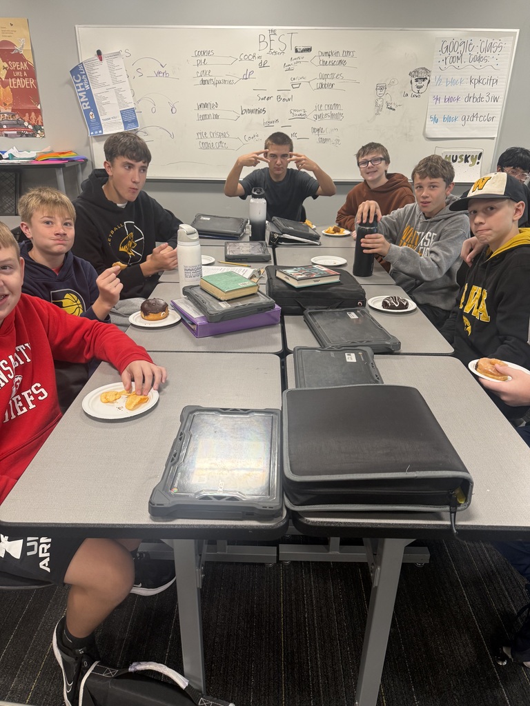 Students seated at a table in a classroom eating lunch and engaging in conversation. Whiteboard and various items on the table.