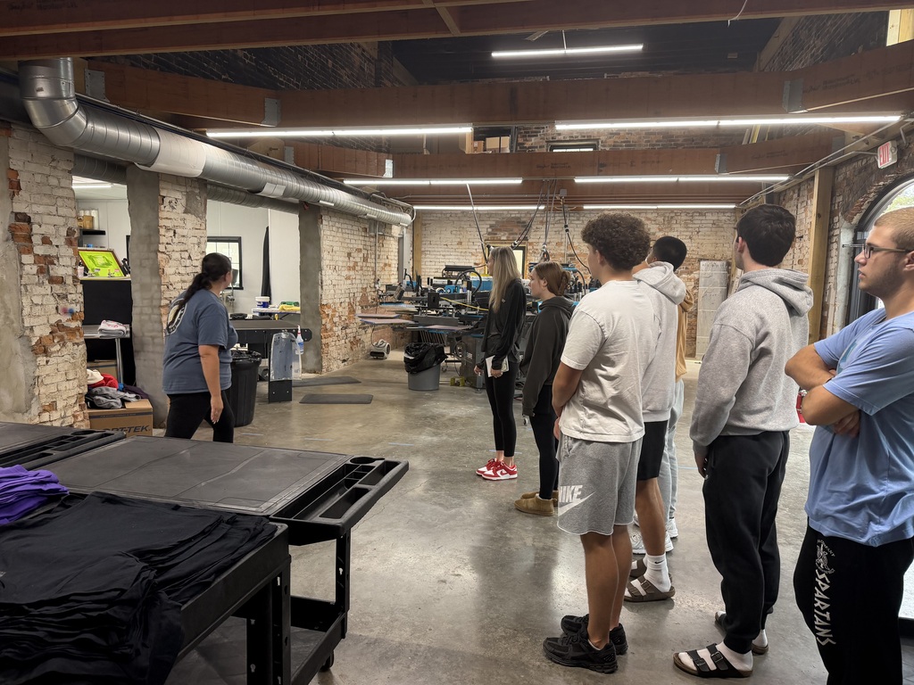 A group of people stand in a room with brick walls, looking at equipment and tables with shirts.