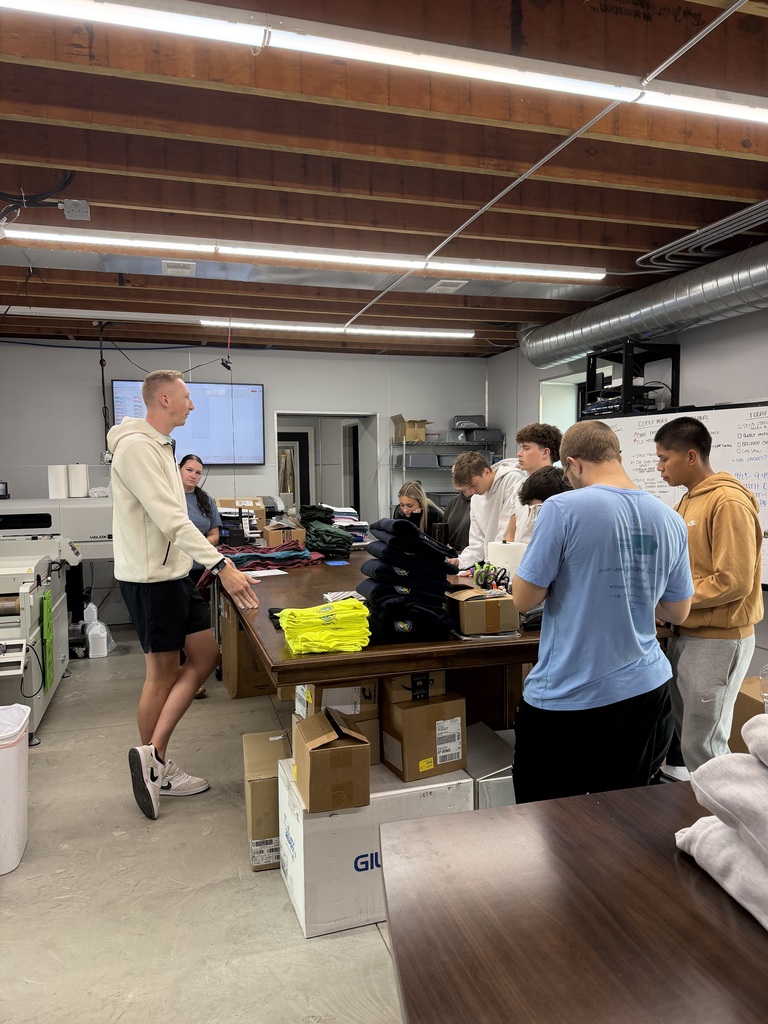 A group of people work around a table in a room. Some stand, others sit. Boxes and bags are on the table.
