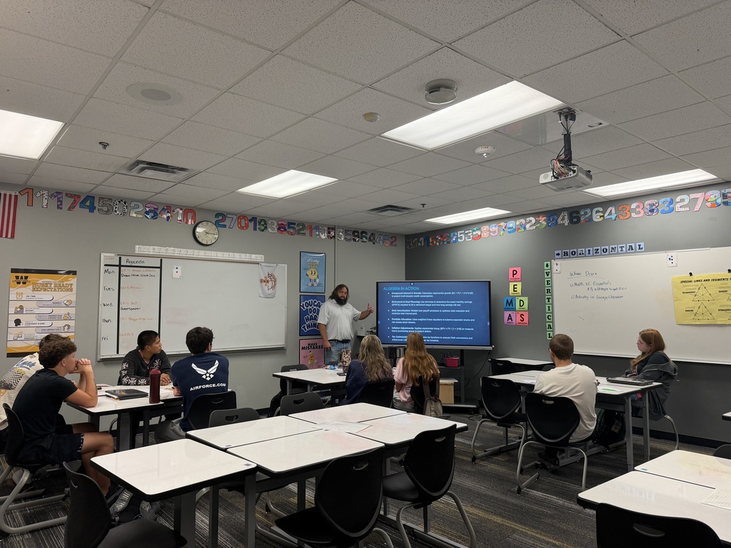 A classroom with students seated around desks, one standing near a projector screen with a presentation.