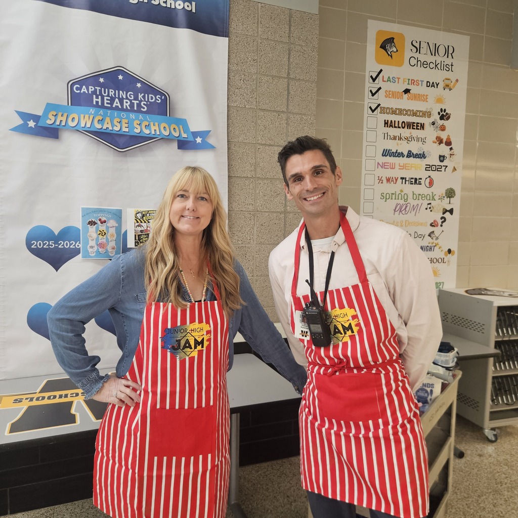 A man and a woman in aprons stand next to a table and banner in a school hallway.