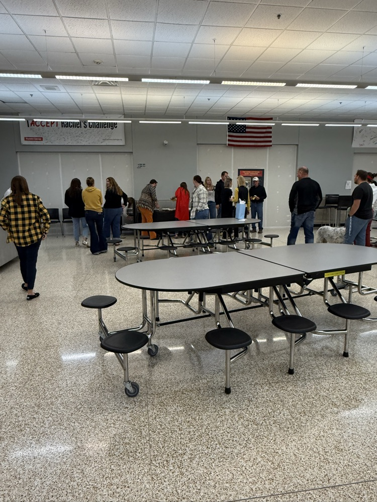Multiple people gather in a cafeteria area with tables and chairs, a flag in the background.