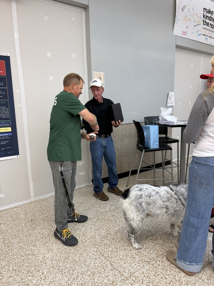Three people, including two men and a woman, are standing in a room with a dog. The man in the green shirt holds a black box.