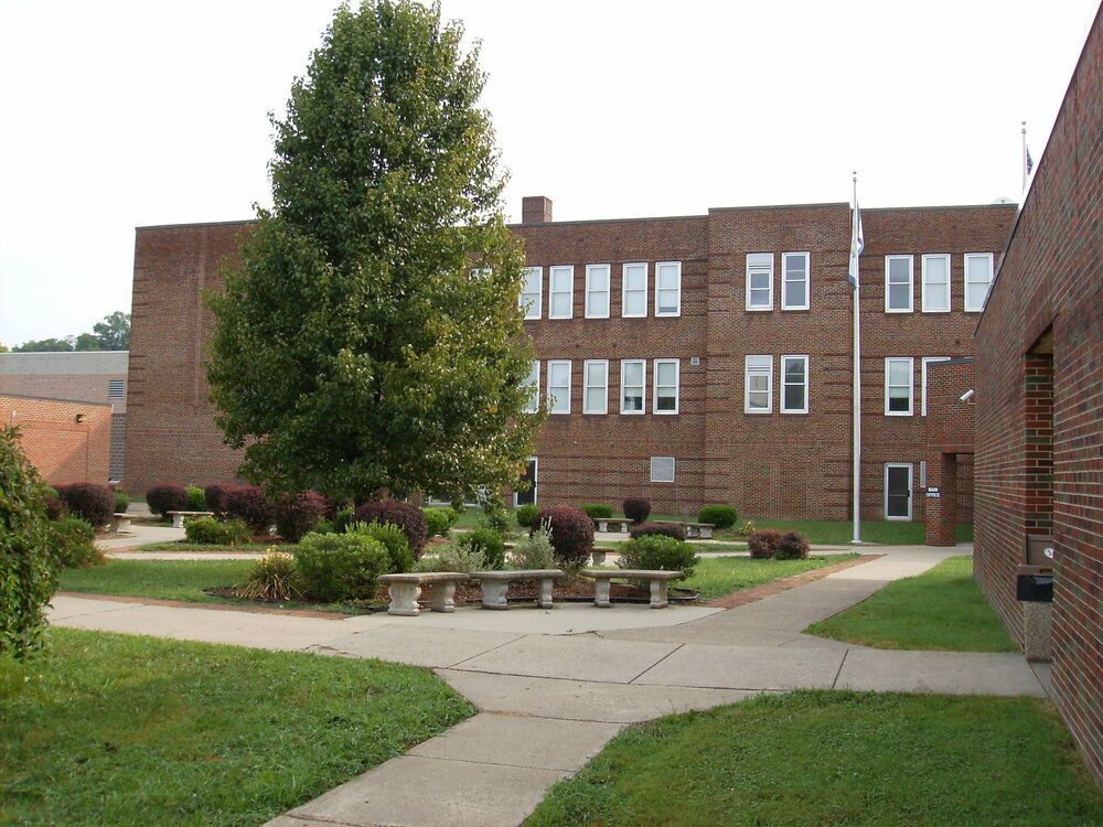Wirt County High School building exterior and lawn. Here are a few concise, accessibility-friendly alt text options you can copy and paste: Short: Exterior view of the Wirt County High School brick building and front lawn. Detailed: Red brick exterior of Wirt County High School with large windows, manicured green lawn, trees, and stone planters in the foreground under a clear sky.