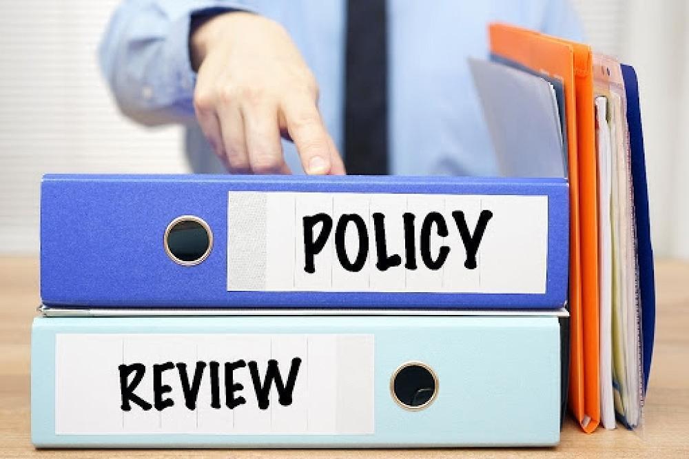 The image shows two stacked office binders labeled "POLICY" (top, blue) and "REVIEW" (bottom, light blue) on a wooden desk.