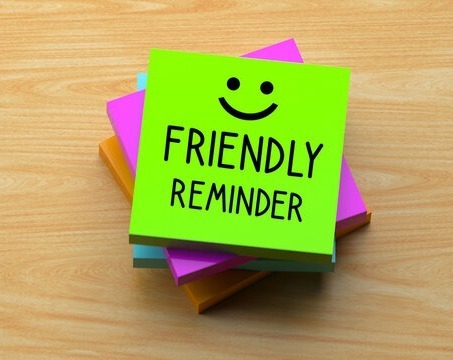 "A stack of colorful sticky notes on a wooden surface, with a bright green sticky note on top that features a hand-drawn smiley face and the text 'FRIENDLY REMINDER'."