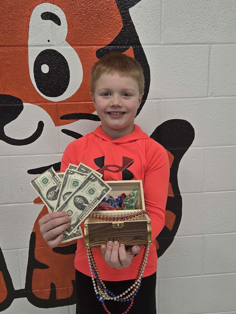 Huntar Kerns wearing a bright red hooded Under Armour shirt is standing in front of a wall mural featuring a large cartoon tiger. The student is holding several one‑dollar bills in one hand and an open wooden treasure chest in the other. The treasure chest contains colorful plastic jewels and beaded necklaces that hang over the front edge.